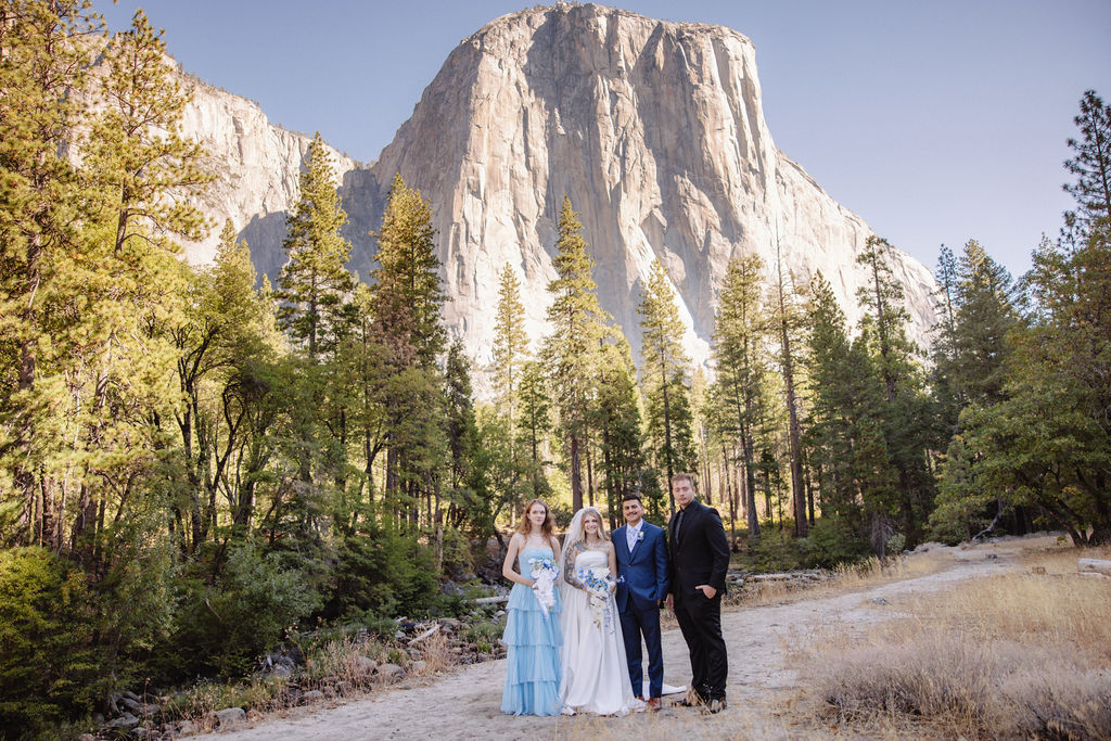 Four people in formal attire stand on a path in a forested area with a large mountain in the background.