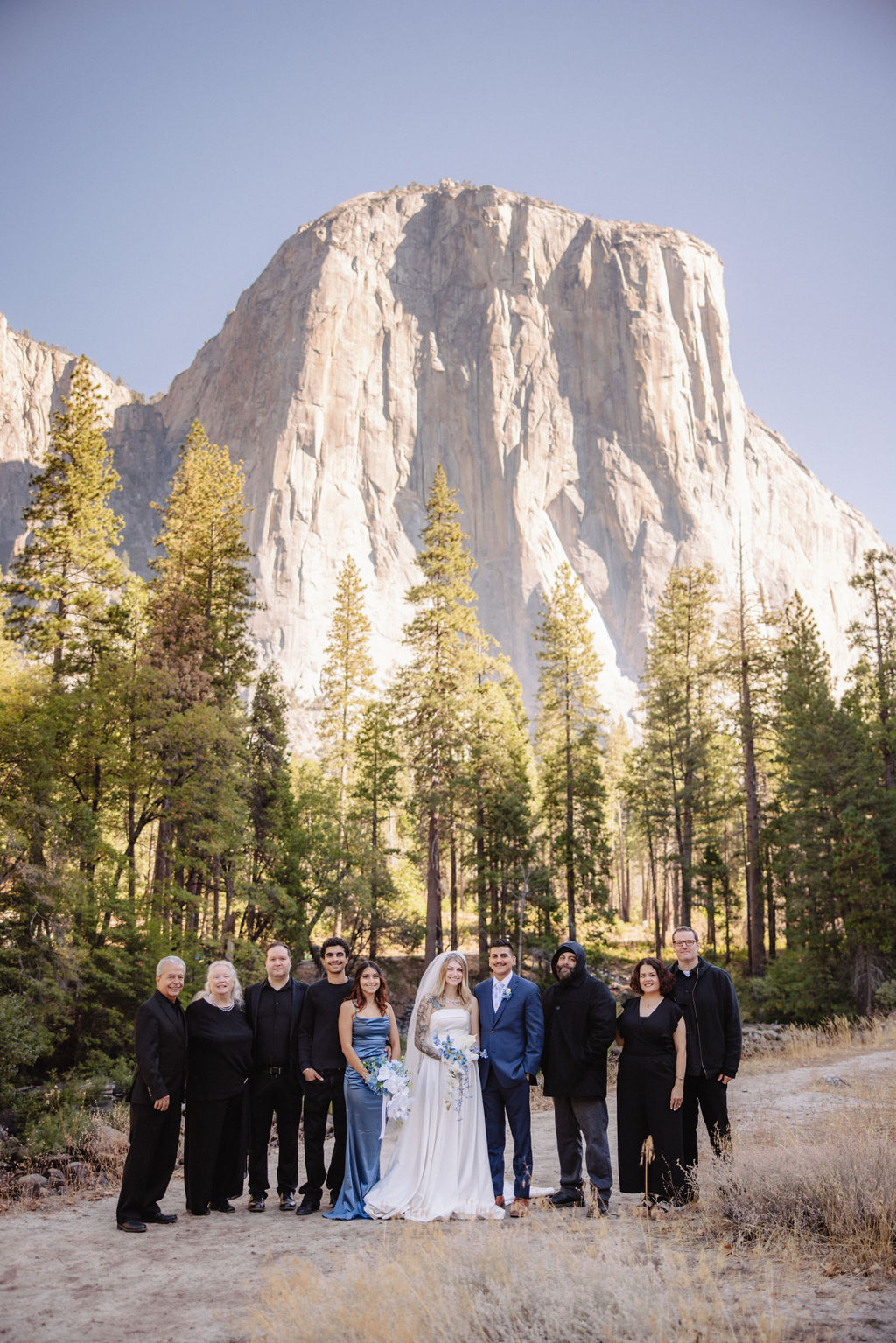 Four people in formal attire stand on a path in a forested area with a large mountain in the background.