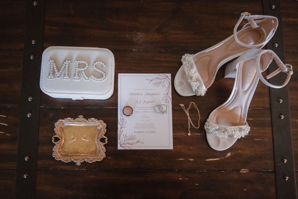 Wedding accessories on a wooden surface: a "Mrs" clutch, high-heeled shoes, invitation card, jewelry, and a decorative box.