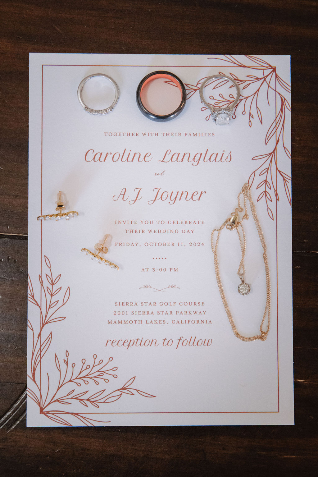 Wedding invitation with gold and red floral design, featuring rings, earrings, and necklace placed on it. Text includes names, date, time, and reception details.