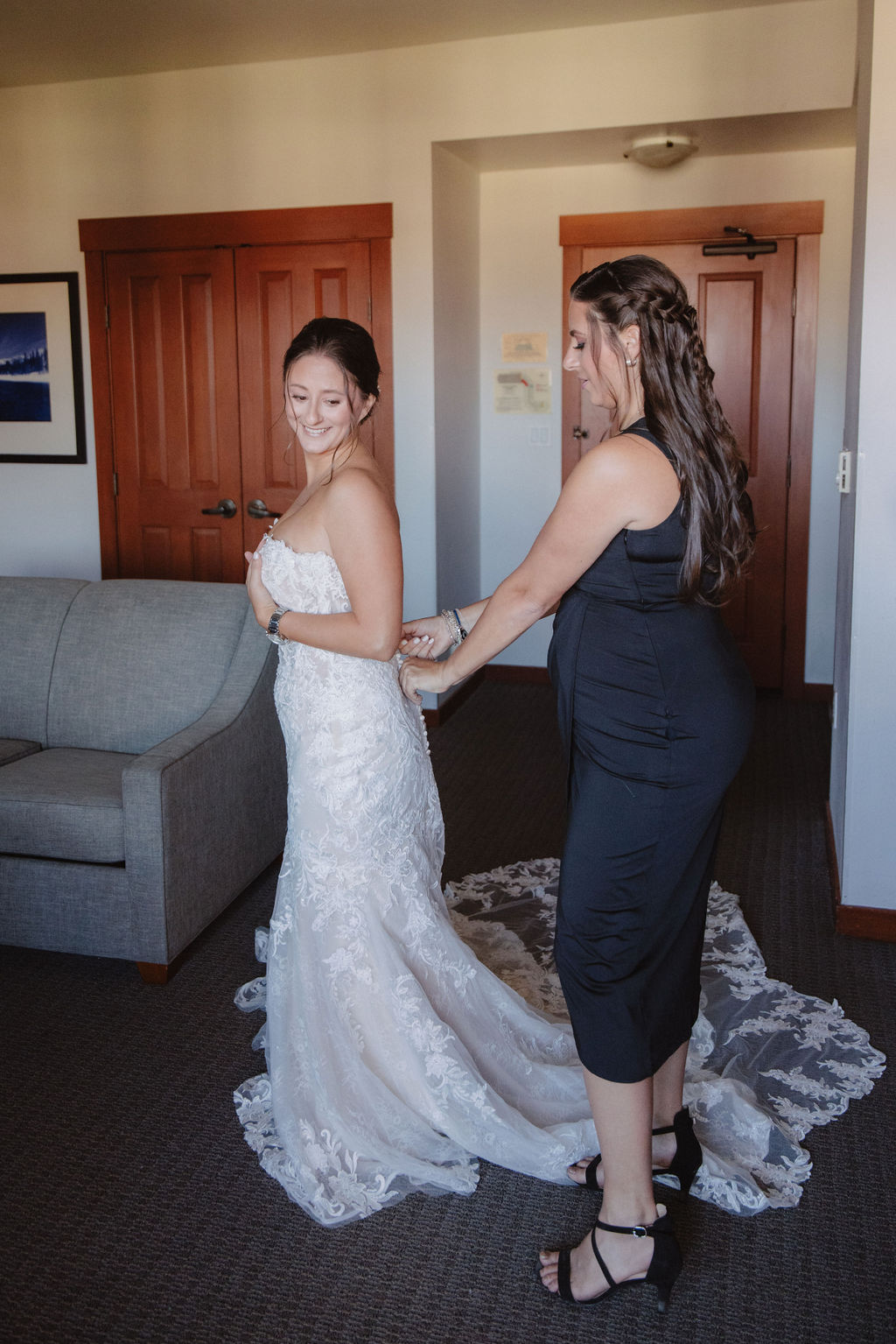 A bride in a white gown is being helped by a woman in a black dress in a room with wooden doors and a gray sofa for a wedding at sierra star golf course