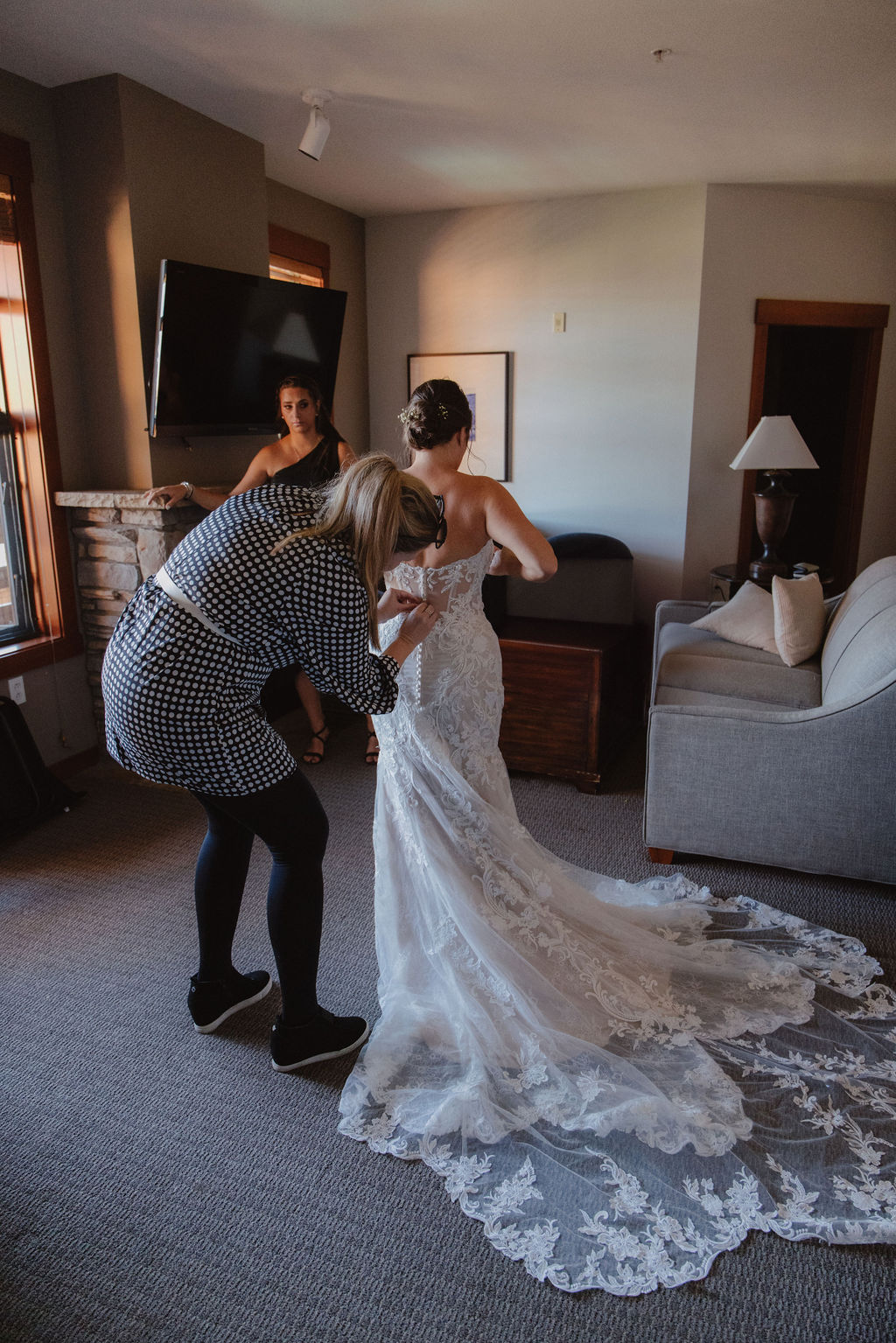 A bride in a white gown is being helped by a woman in a black dress in a room with wooden doors and a gray sofa for a wedding at sierra star golf course