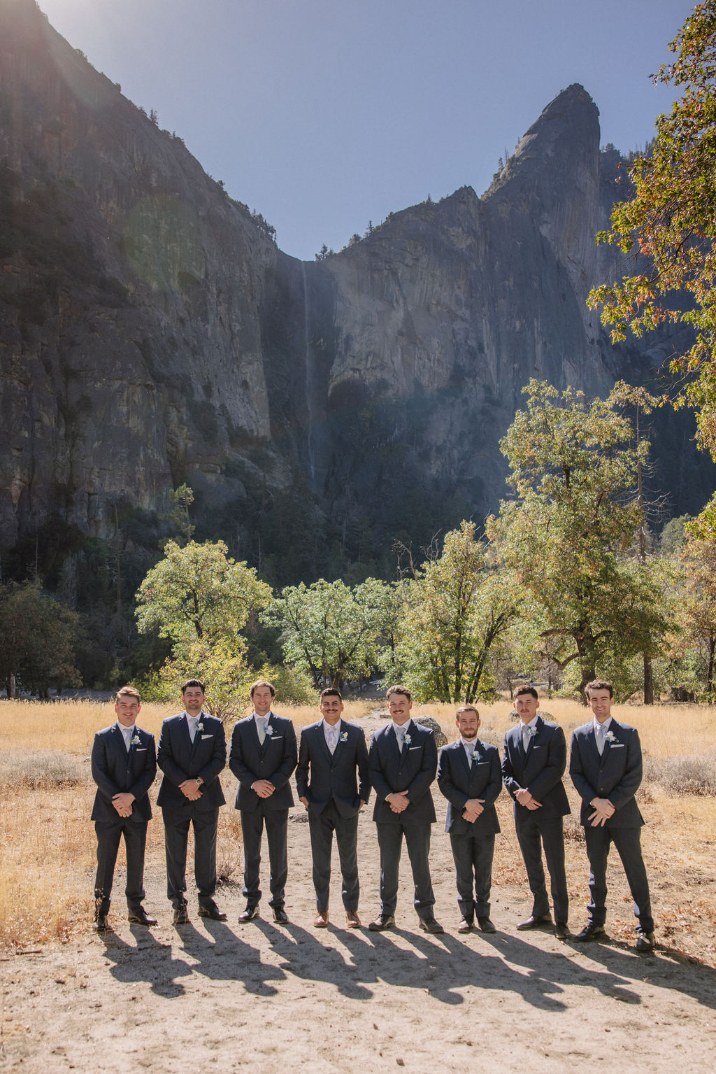 A group of men in suits poses playfully in a sunlit field with mountains in the background.