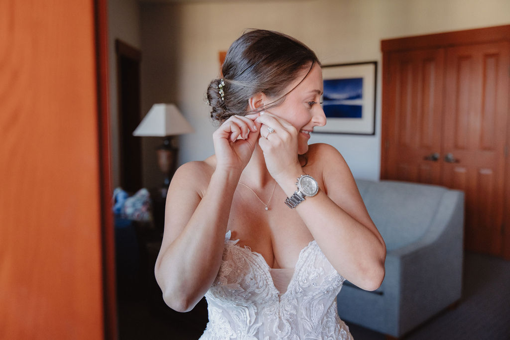 A woman in a white dress stands in a room, smiling as she adjusts her earring for a wedding at sierra star golf course