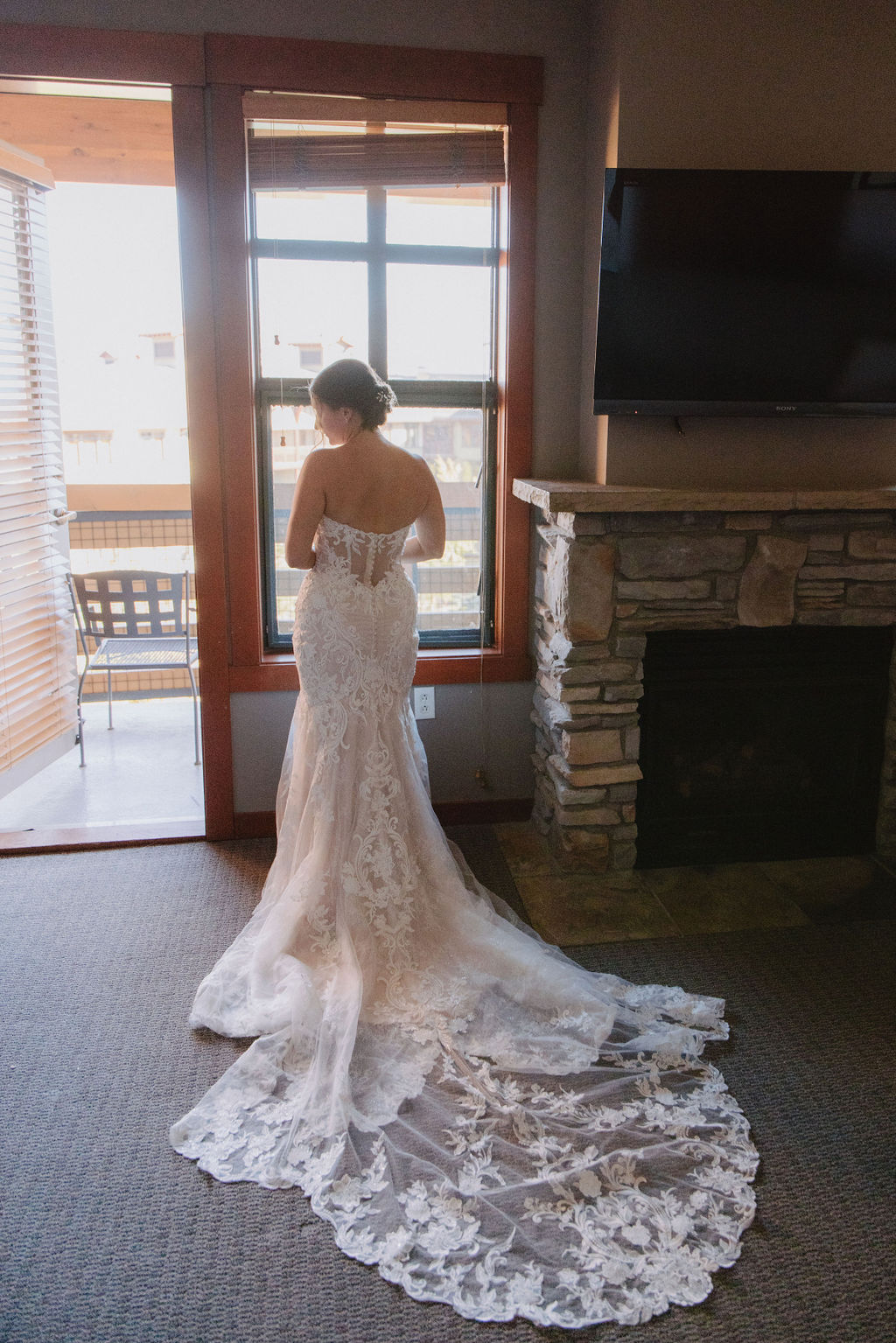 A woman in a white lace wedding gown stands facing a window beside a stone fireplace, with the gown's train spread out on the floor.