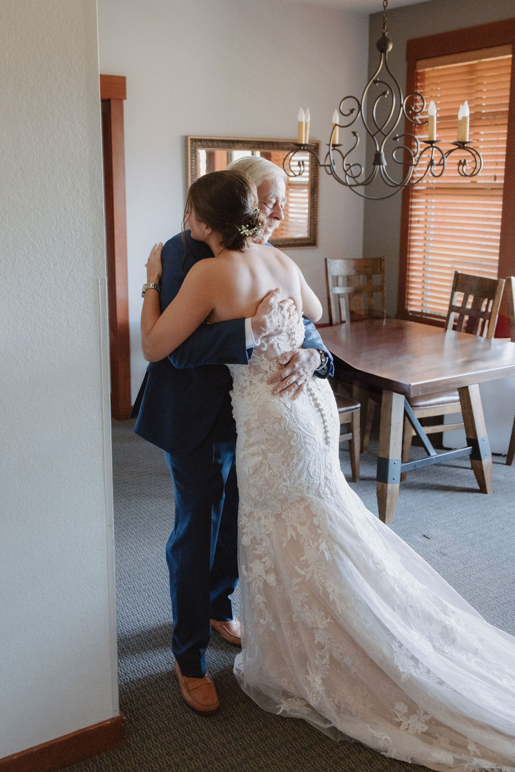 A bride in a white gown hugs an older man in a suit inside a room with a wooden table and chandelier.