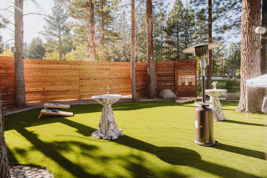 Outdoor event space with high tables on artificial grass, surrounded by trees and a wooden fence. A heater and cornhole game set are also visible.