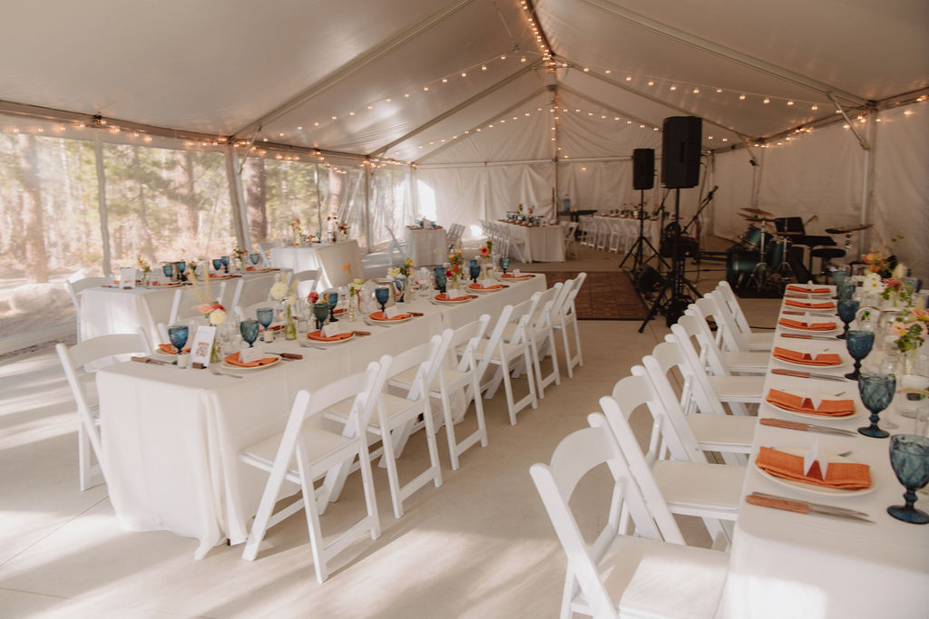 A decorated event tent with tables set for a formal gathering. Tables have white cloths, orange napkins, and blue glasses. White chairs surround the tables, and string lights hang above at a wedding at sierra star golf course