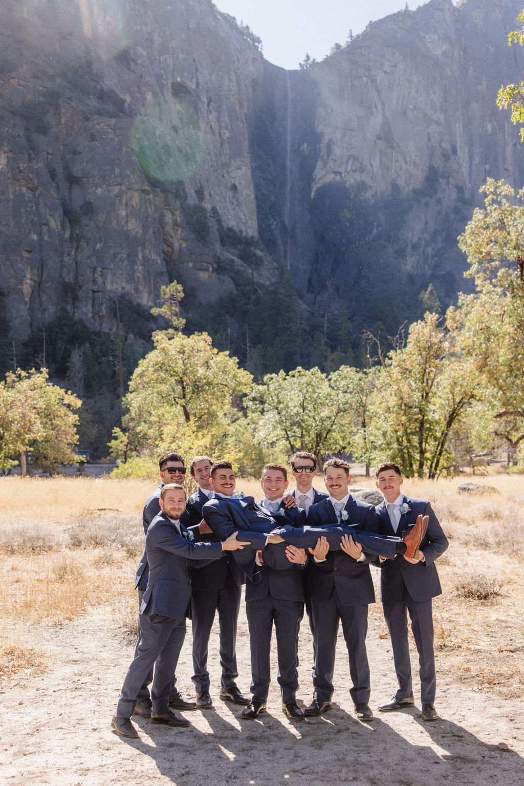 A group of men in suits poses playfully in a sunlit field with mountains in the background.