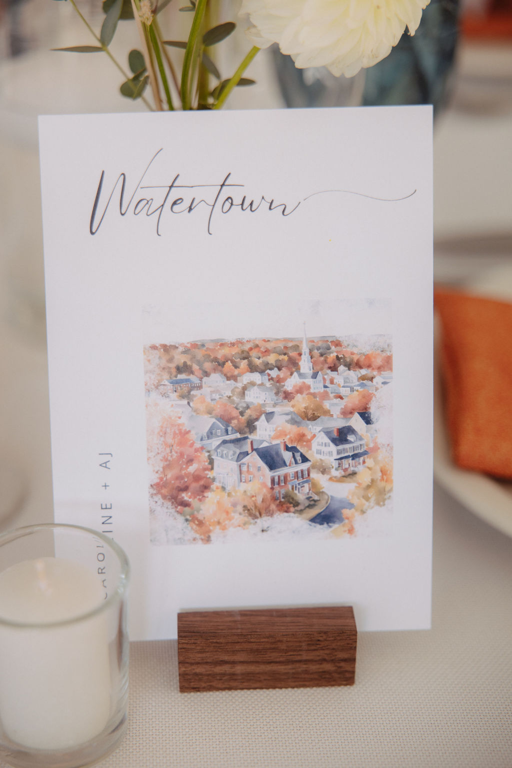 A card labeled "Watertown" with an image of autumn-colored trees and houses is displayed on a wooden holder. A small candle and an orange napkin are nearby.