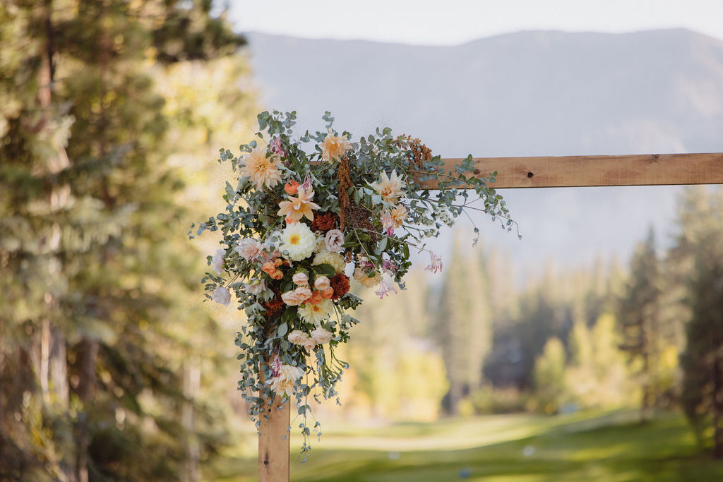 A wooden wedding arch decorated with flowers stands on a grassy area surrounded by tall trees, with a microphone setup in the center. A mountain range is visible in the background at a wedding at sierra star golf course