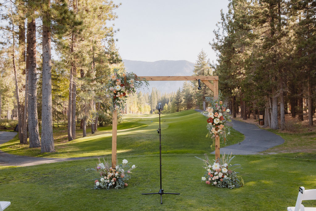 A wooden wedding arch decorated with flowers stands on a grassy area surrounded by tall trees, with a microphone setup in the center. A mountain range is visible in the background at a wedding at sierra star golf course