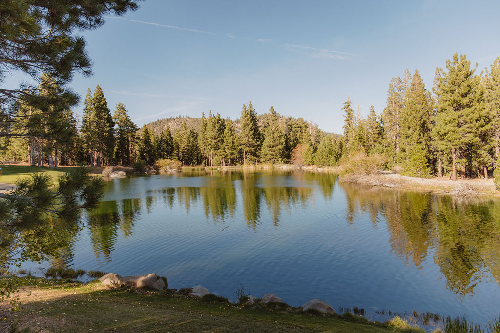 A tranquil lake reflecting surrounding pine trees and a clear blue sky, with forested hills in the background for a wedding at sierra star golf course