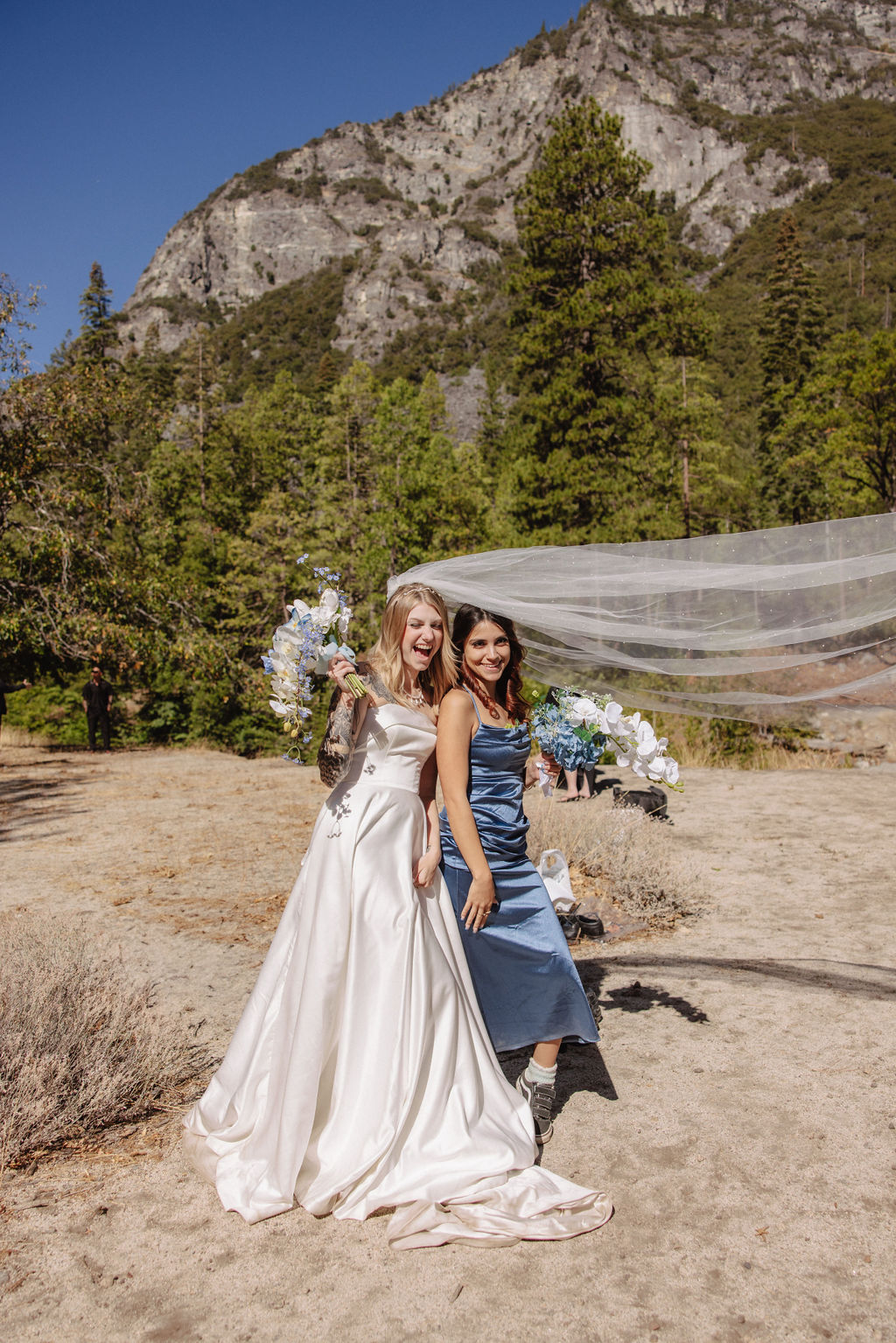 A bride in a white gown and a woman in a blue dress stand outdoors with a mountainous backdrop.