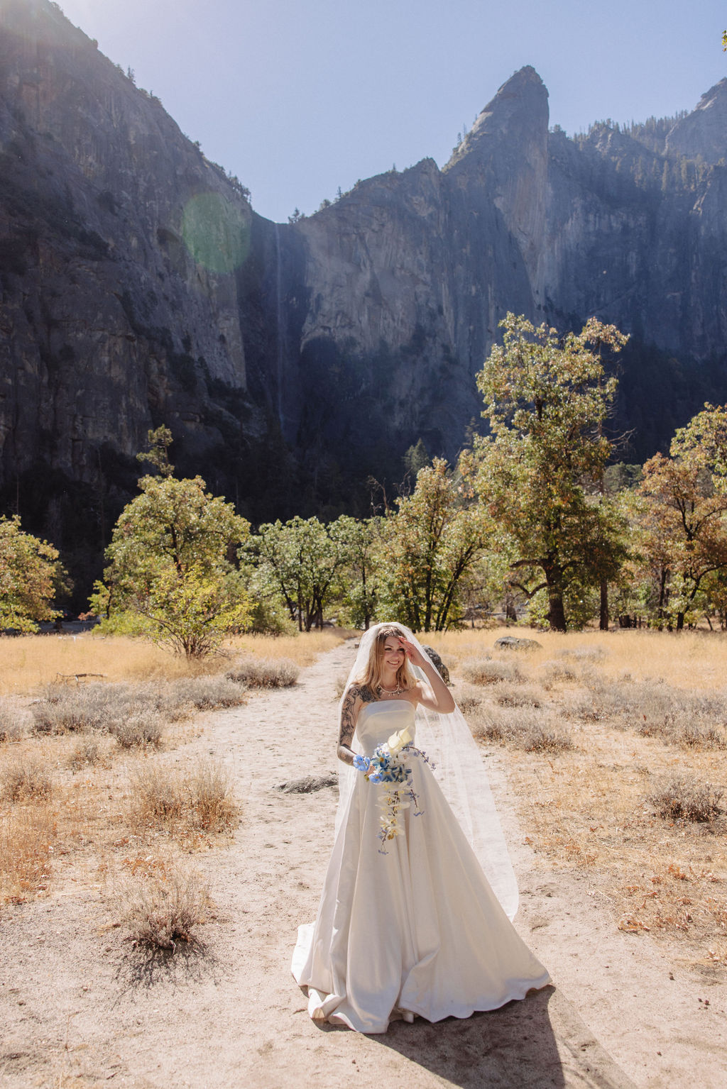 Bride in a strapless white dress and veil stands holding a bouquet in a sunlit, rocky landscape with mountains and sparse trees in the background.