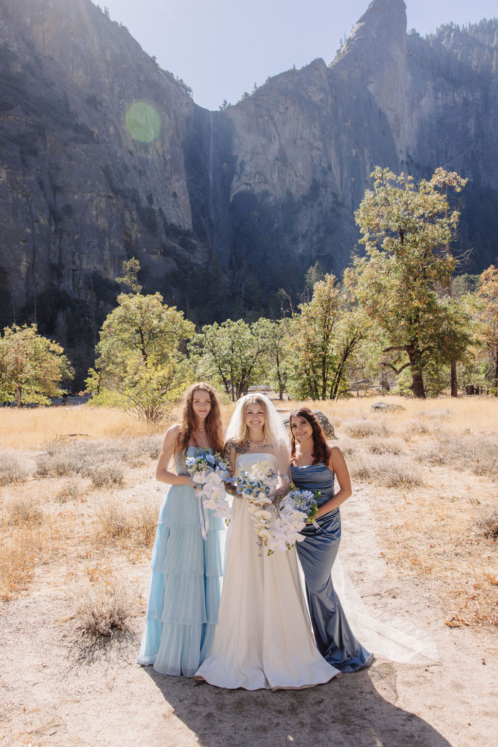 Three women in formal dresses, one in a bridal gown, pose outdoors with a mountain backdrop.