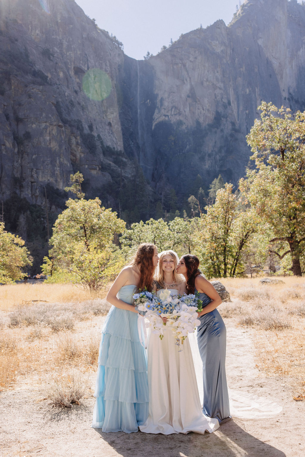 Three women in formal dresses, one in a bridal gown, pose outdoors with a mountain backdrop.