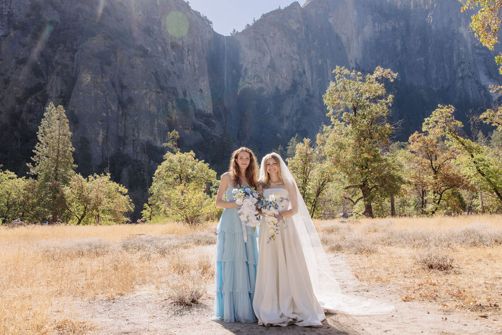 Three women in formal dresses, one in a bridal gown, pose outdoors with a mountain backdrop.