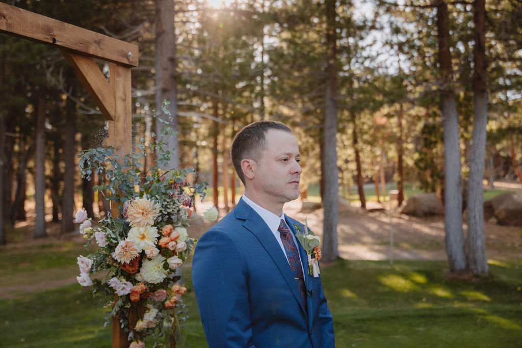 A man in a blue suit stands outdoors near a wooden structure and flower arrangement, surrounded by trees and grass.