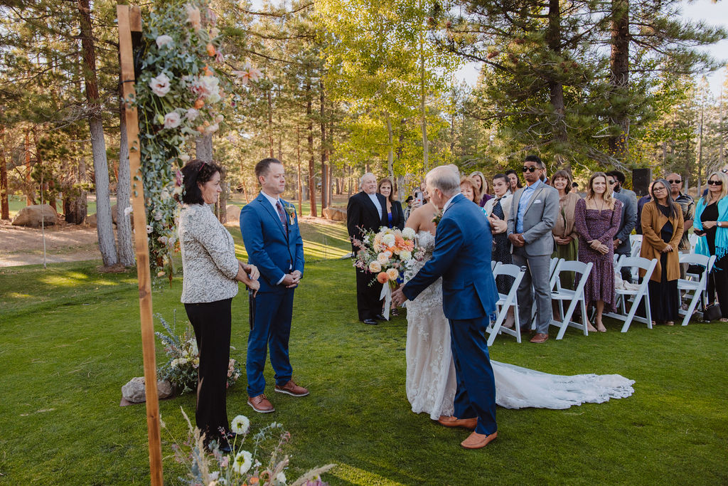 An outdoor wedding ceremony with a bride and groom standing before an officiant. Guests are seated and standing, surrounded by trees.