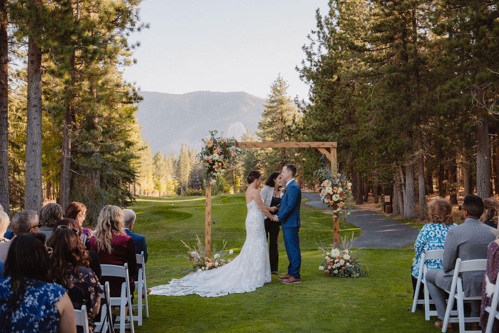 A couple is getting married outdoors under a wooden arch with floral decorations, surrounded by seated guests on a grass lawn with trees and mountains in the background for a wedding at sierra star golf course