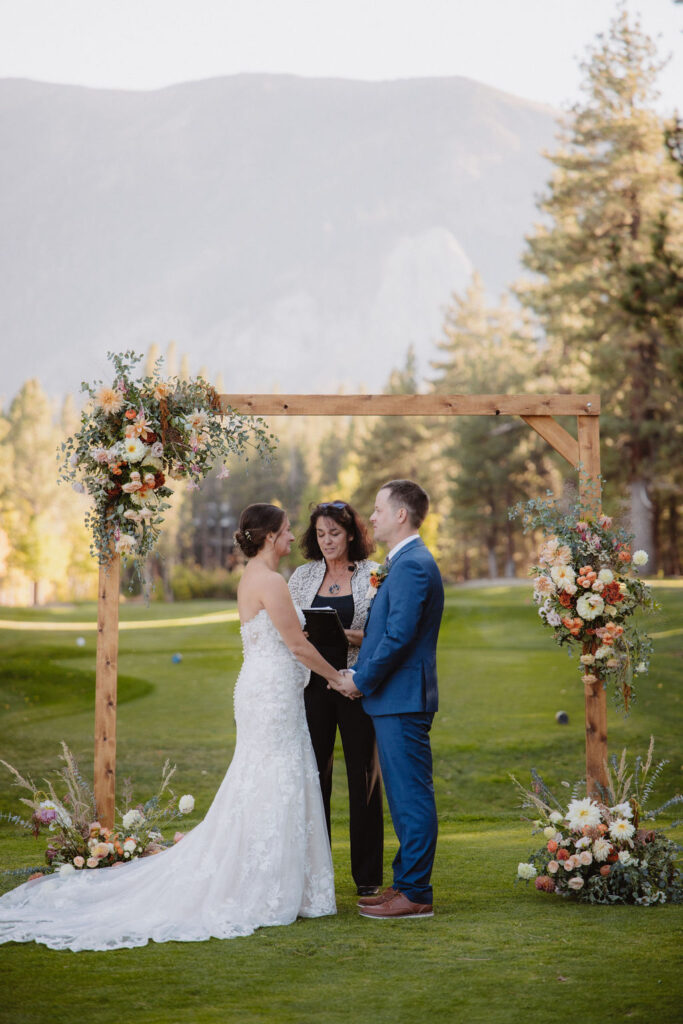 A couple stands holding hands during an outdoor wedding ceremony, with a wooden arch and floral decorations in the backdrop of a grassy area and mountain scenery.