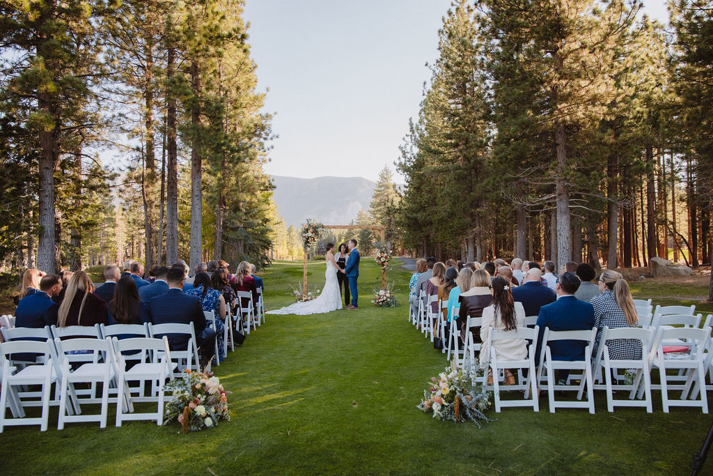 A couple is getting married outdoors under a wooden arch with floral decorations, surrounded by seated guests on a grass lawn with trees and mountains in the background.