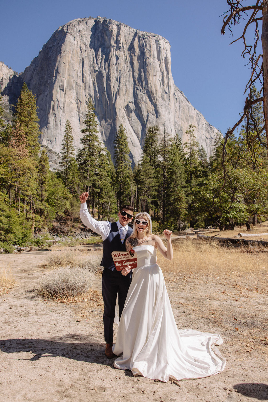 A couple in wedding attire poses with a pizza box in front of a large rock formation and trees under a clear blue sky.