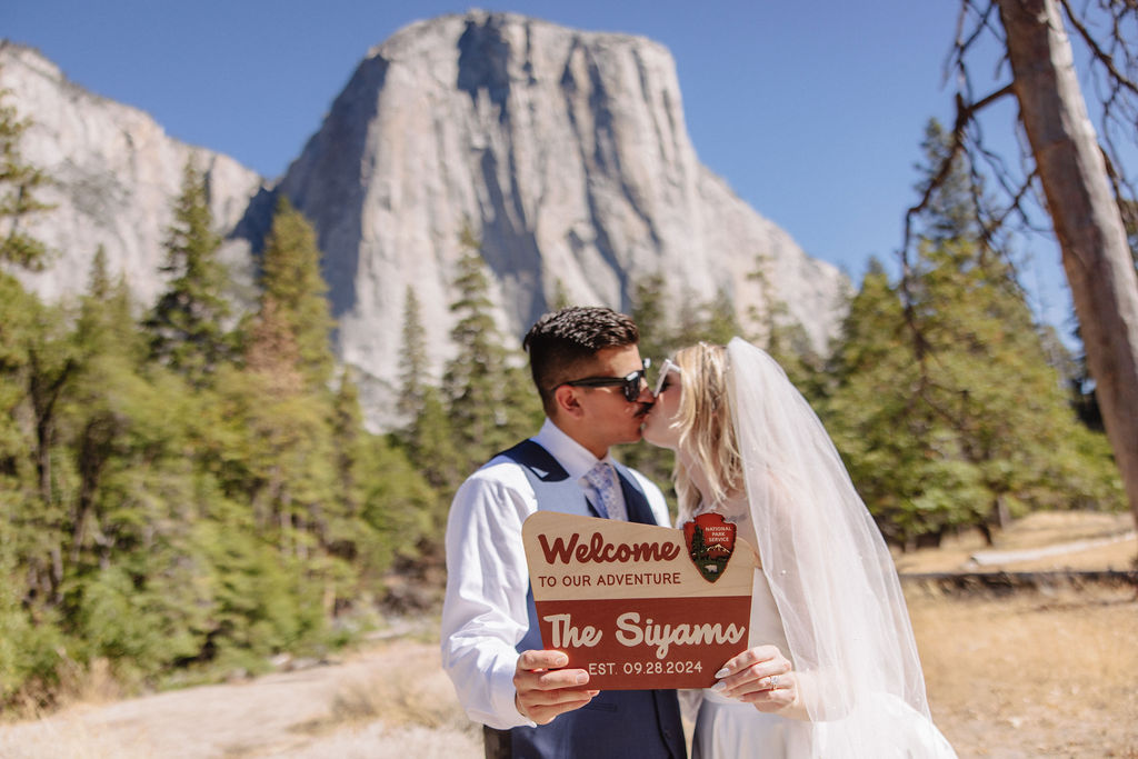 A couple in wedding attire kisses, holding a sign that says, "Welcome to our adventure, The Suyams, October 13th, 2023," with a mountainous backdrop and trees.