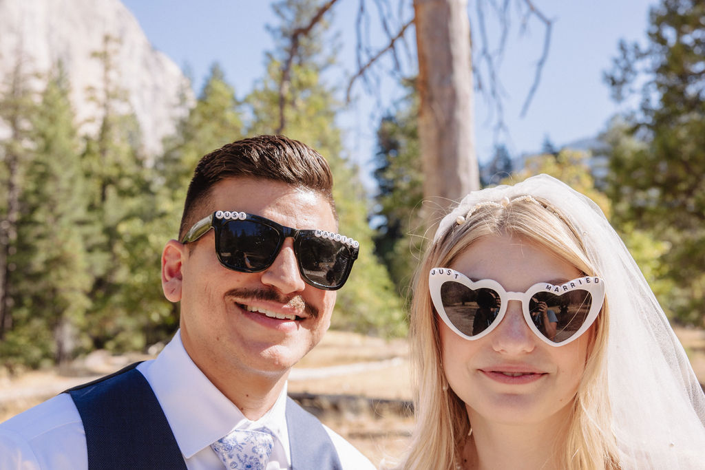 A couple wearing heart-shaped sunglasses smiles outdoors, surrounded by trees and mountains.