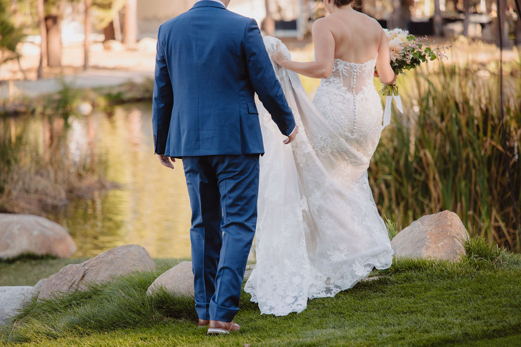 A couple in wedding attire walks near a pond. The groom is in a blue suit, and the bride wears a white dress with a long train.