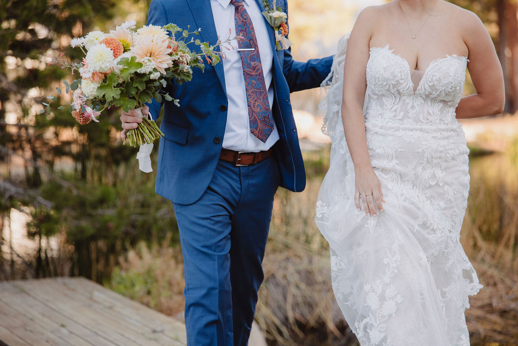 A couple in wedding attire walks near a pond. The groom is in a blue suit, and the bride wears a white dress with a long train.