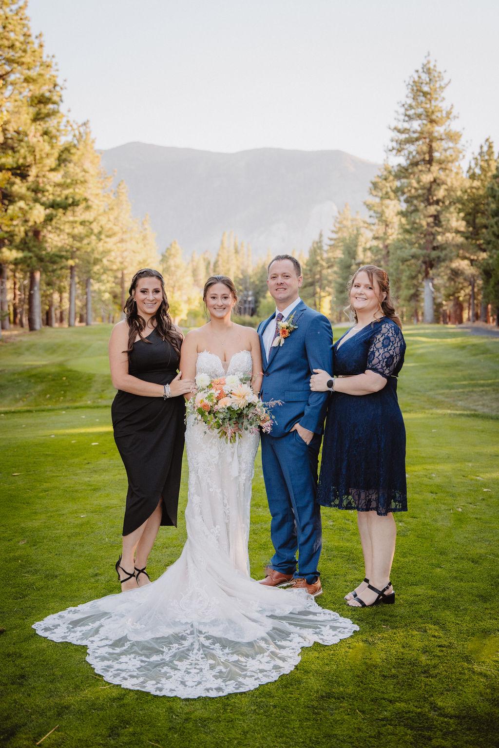 A bride and groom stand outdoors with family members, all dressed in formal attire, surrounded by trees and mountains in the background.