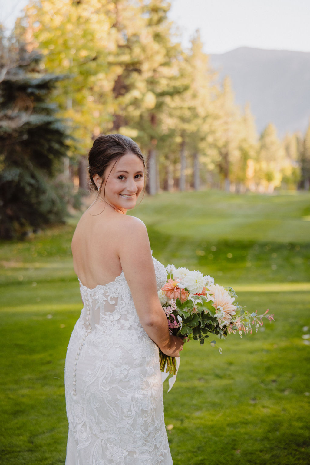 A bride in a strapless lace wedding dress holds a bouquet while standing on a green lawn with trees and mountains in the background.