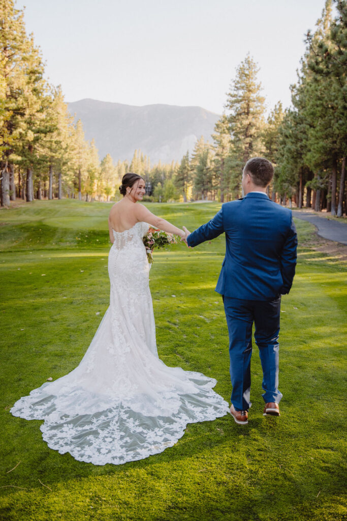 A couple in formal attire holds hands, walking down a grassy path lined with trees and mountains in the background for their wedding at sierra dreamy star golf course