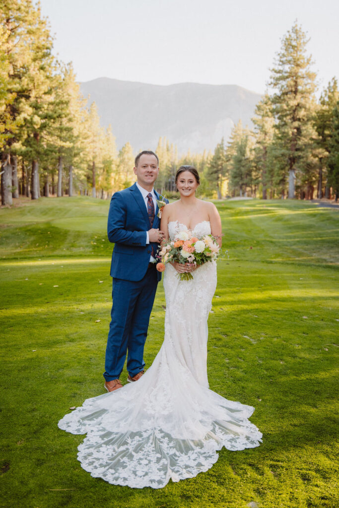 A couple in formal attire holds hands, walking down a grassy path lined with trees and mountains in the background for their wedding at sierra dreamy star golf course