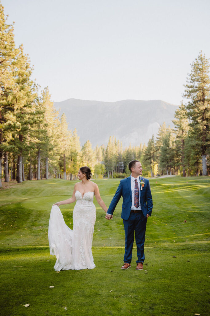 A couple in formal attire holds hands, walking down a grassy path lined with trees and mountains in the background for their wedding at sierra dreamy star golf course