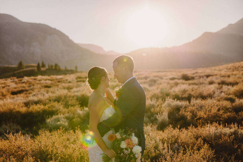 A couple embraces outdoors during sunset, with the sun shining in the background and mountains in the distance. The person in a wedding dress is seen from behindnfor their wedding at Sierra Star Golf Course