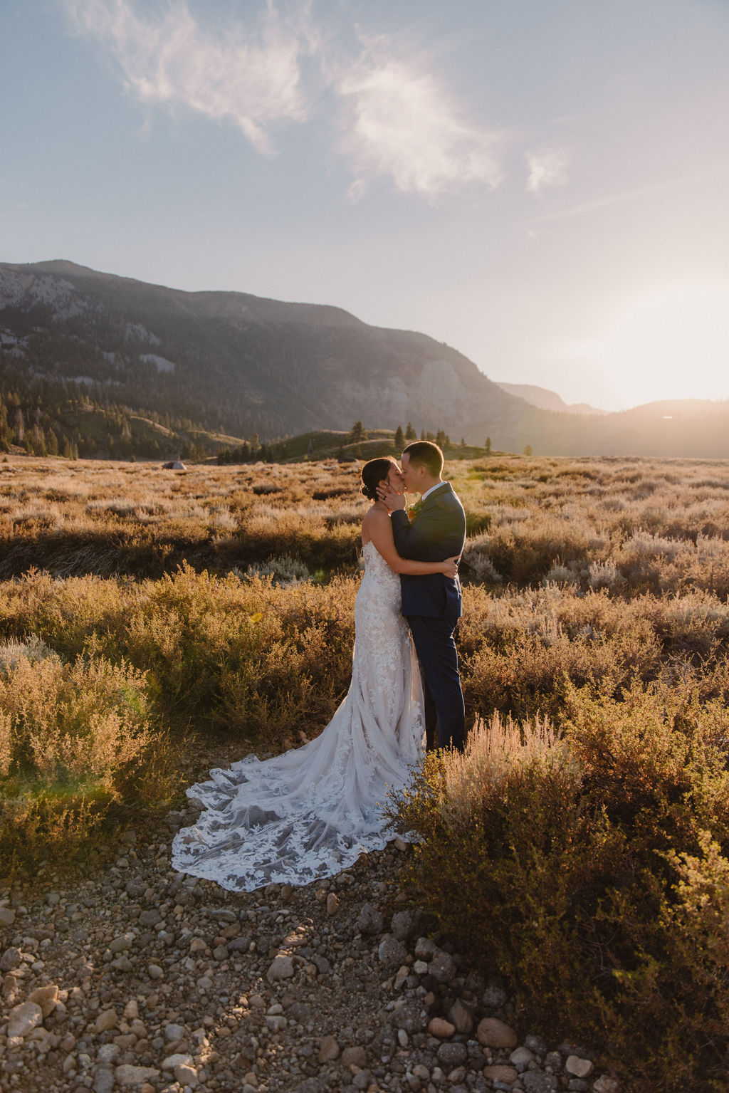 A couple embraces outdoors during sunset, with the sun shining in the background and mountains in the distance. The person in a wedding dress is seen from behindnfor their wedding at Sierra Star Golf Course