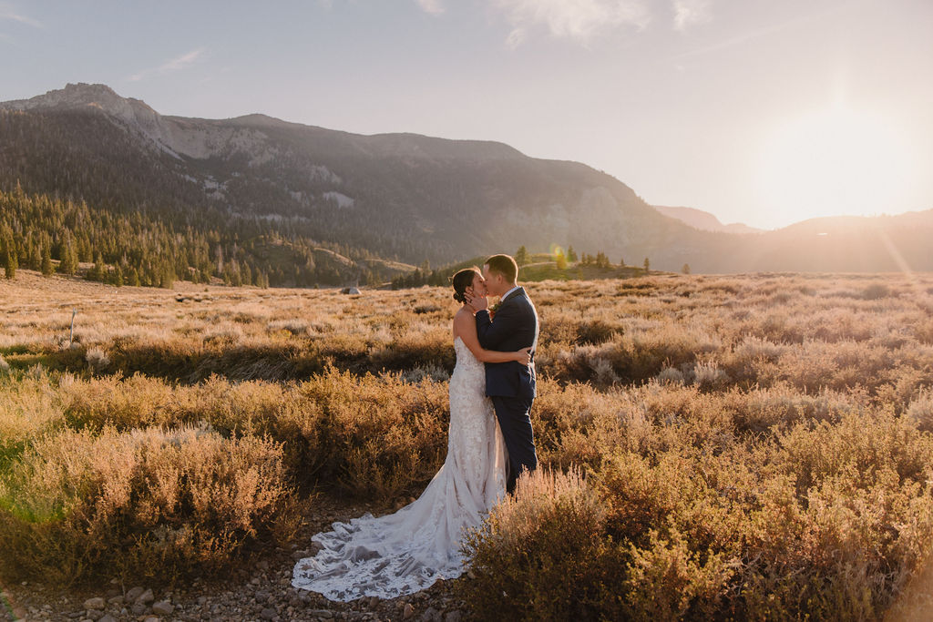 A couple embraces outdoors during sunset, with the sun shining in the background and mountains in the distance. The person in a wedding dress is seen from behindnfor their wedding at Sierra Star Golf Course