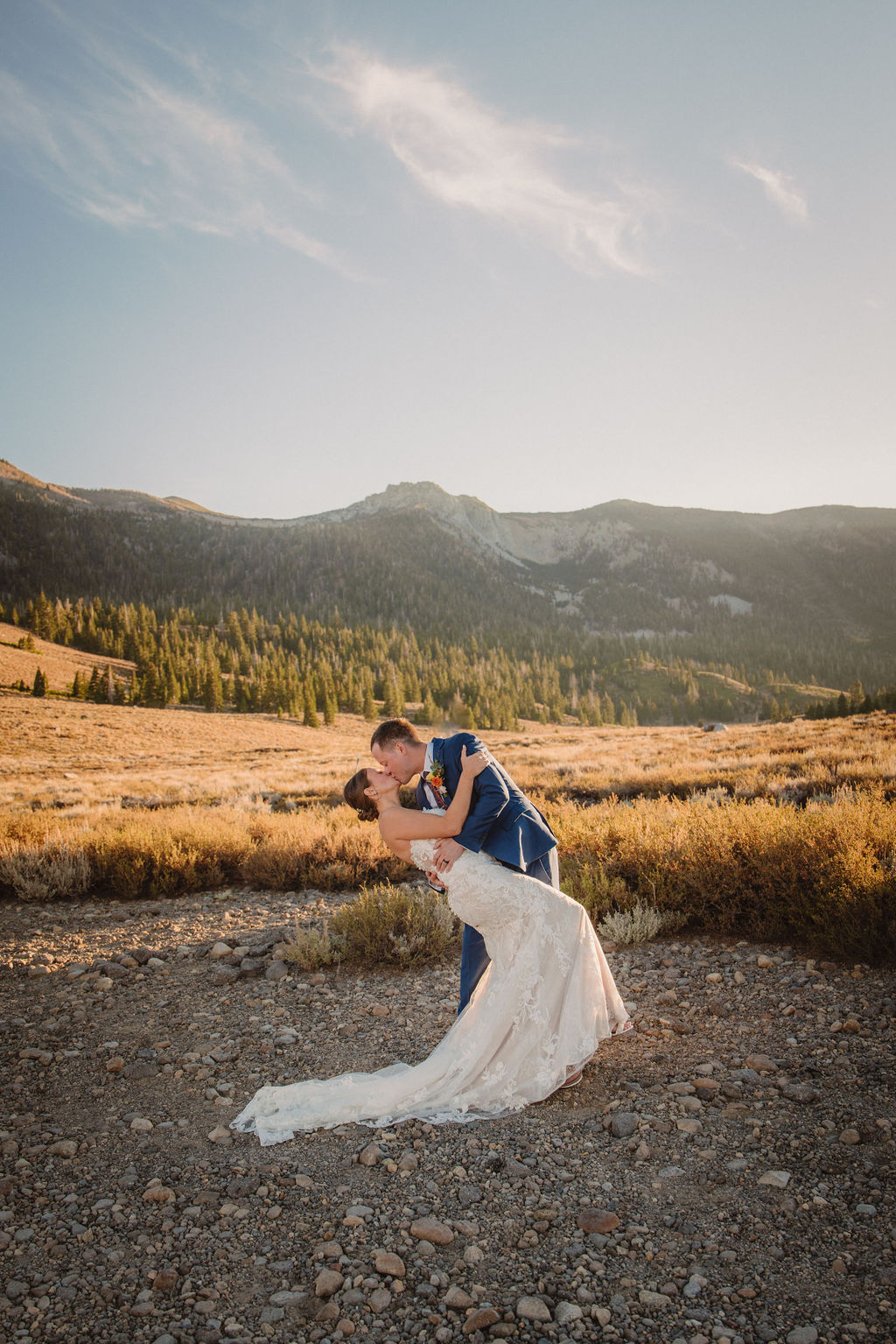 A couple embraces outdoors during sunset, with the sun shining in the background and mountains in the distance. The person in a wedding dress is seen from behindnfor their wedding at Sierra Star Golf Course