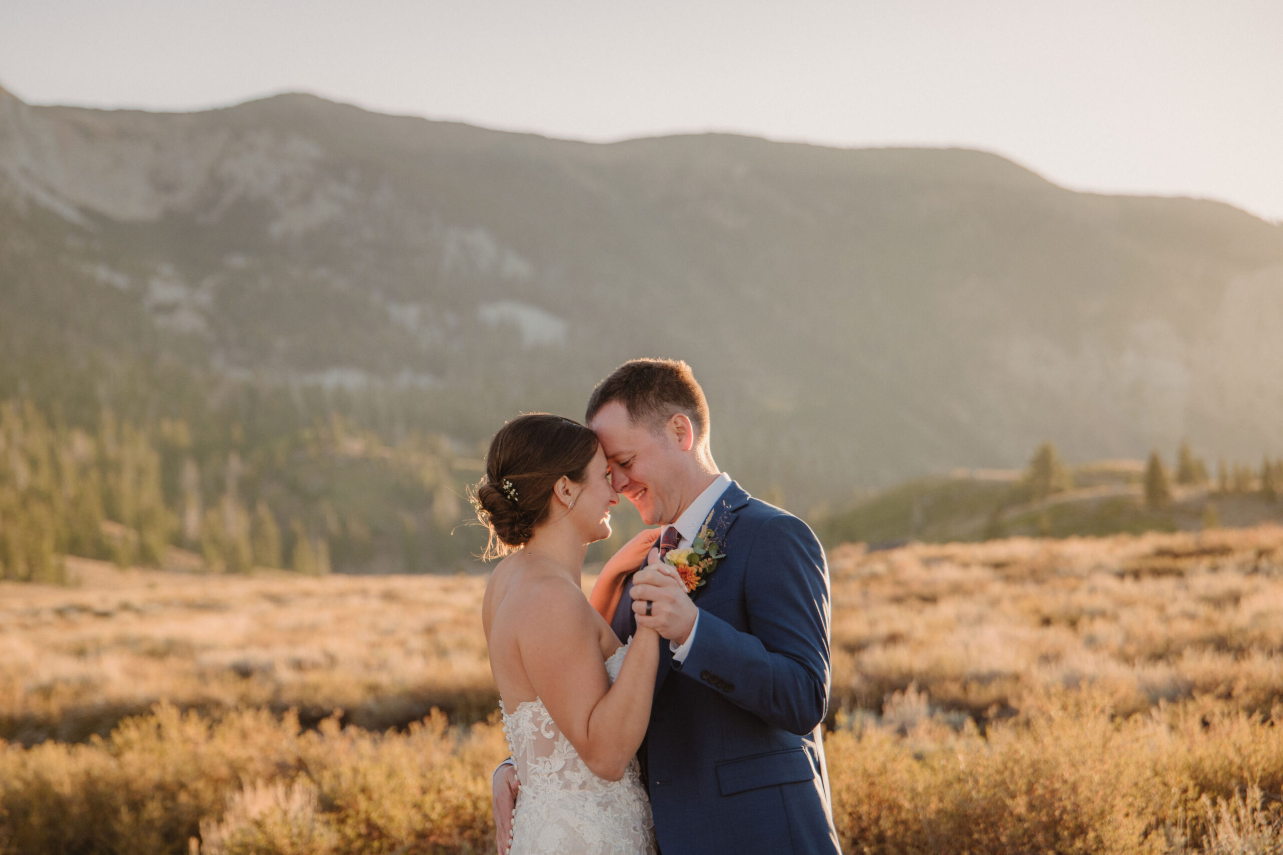 A bride in a strapless lace wedding dress holds a bouquet while standing on a green lawn with trees and mountains in the background.