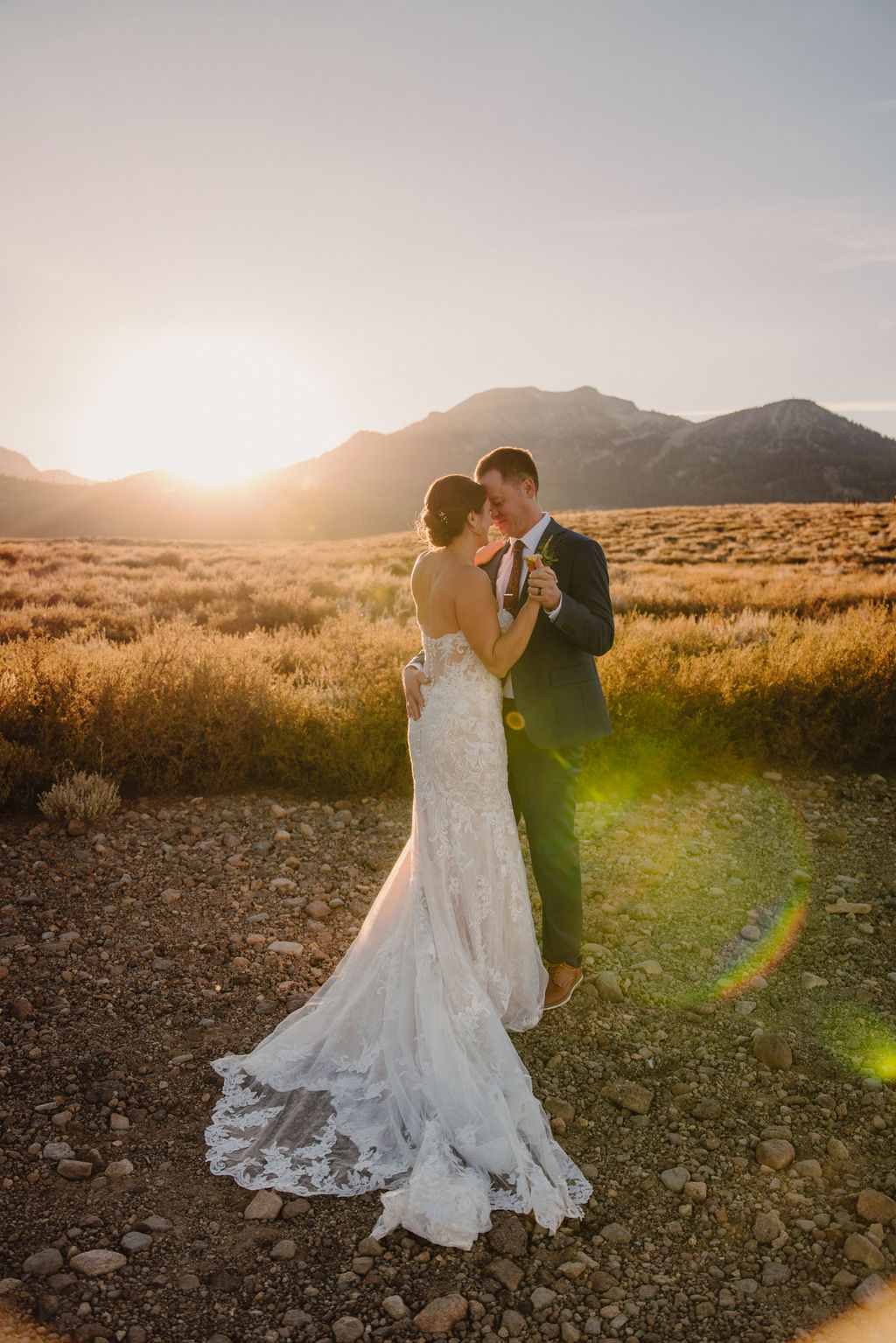 A couple embraces outdoors during sunset, with the sun shining in the background and mountains in the distance. The person in a wedding dress is seen from behindnfor their wedding at Sierra Star Golf Course