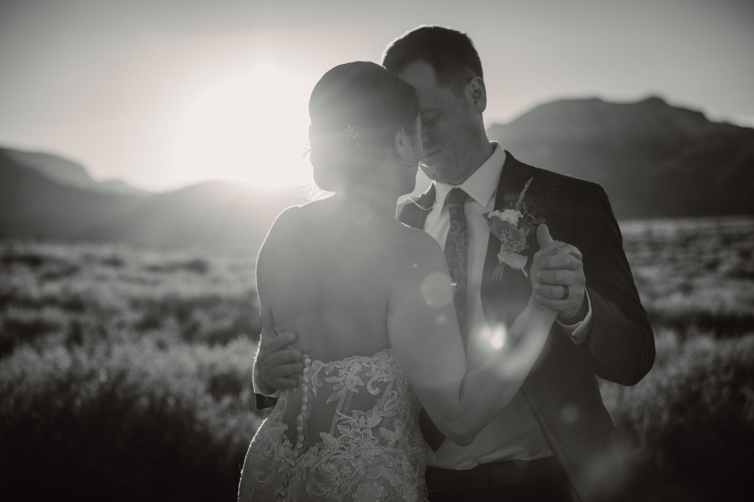 A bride in a strapless lace wedding dress holds a bouquet while standing on a green lawn with trees and mountains in the background.