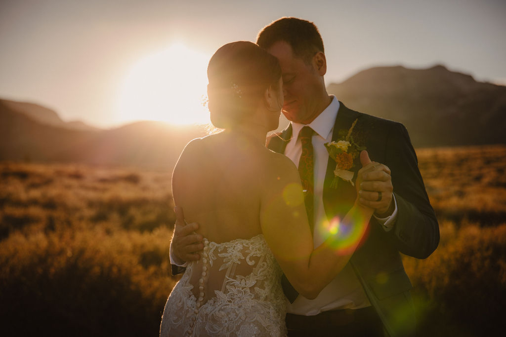 A couple embraces outdoors during sunset, with the sun shining in the background and mountains in the distance. The person in a wedding dress is seen from behindnfor their wedding at Sierra Star Golf Course