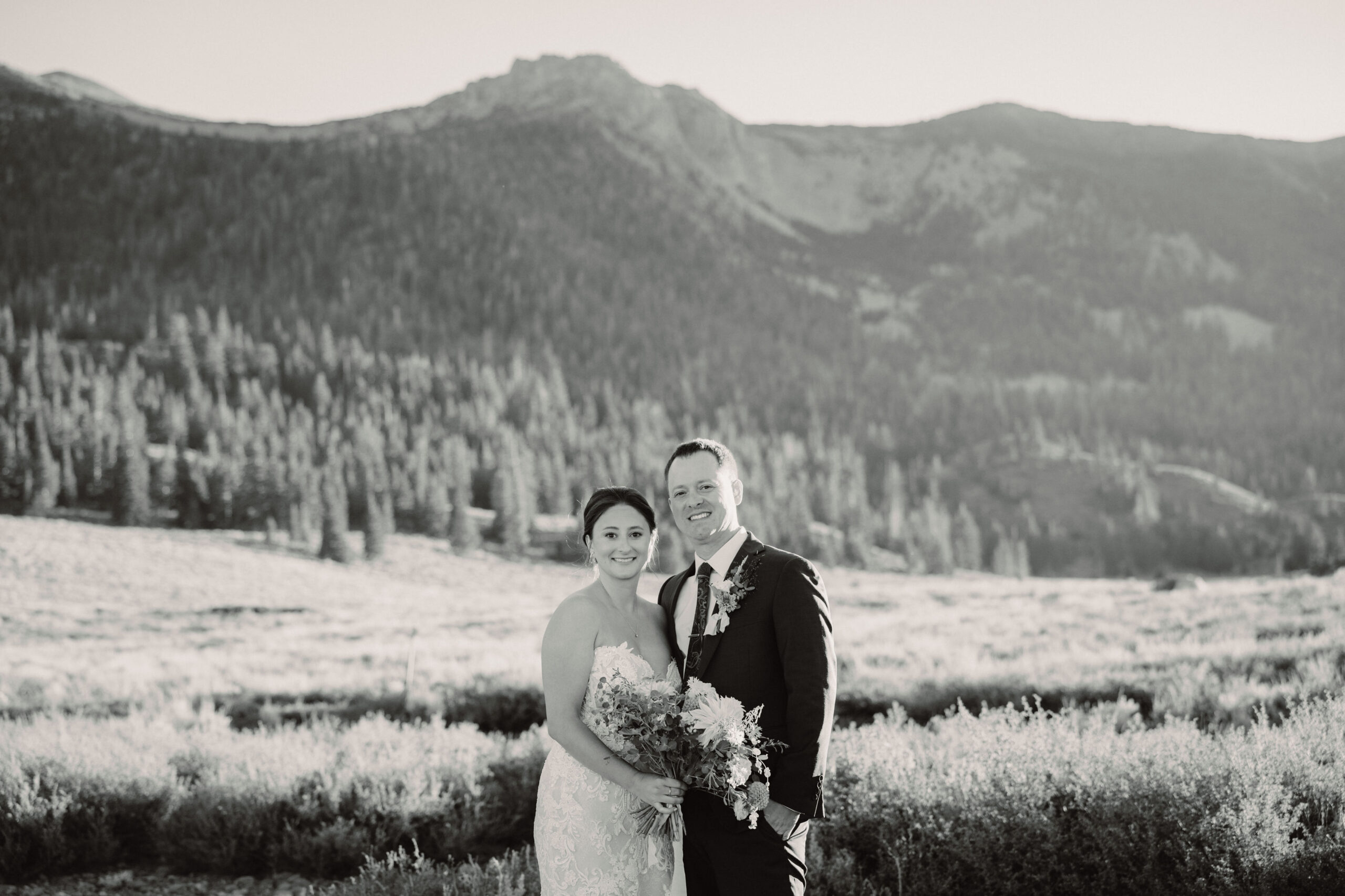 A bride in a strapless lace wedding dress holds a bouquet while standing on a green lawn with trees and mountains in the background.