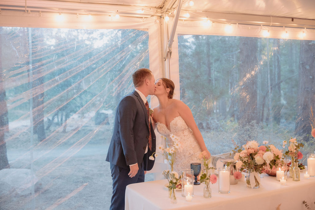 A couple in wedding attire kisses by a decorated table under string lights in a tent. at a wedding at sierra star golf course