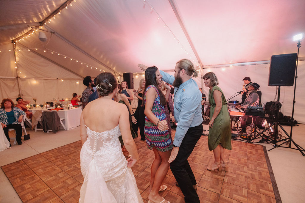 People dancing on a wooden floor under a tent with string lights, while a band plays in the background.