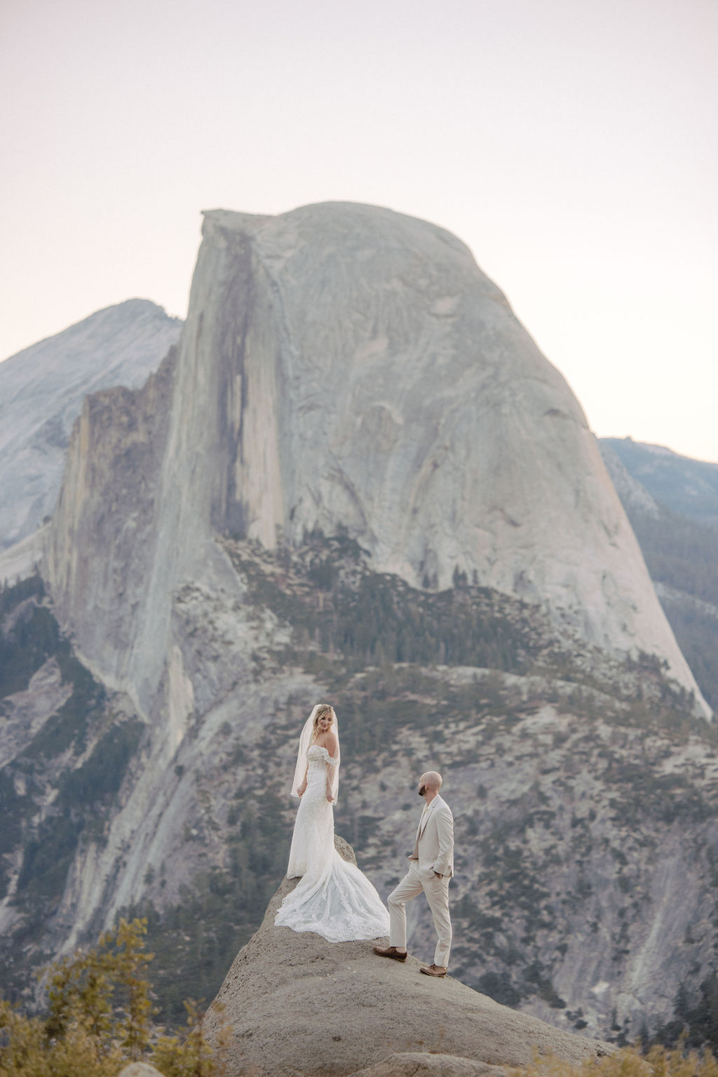 A bride and groom stand on a rock with a mountain landscape in the background.