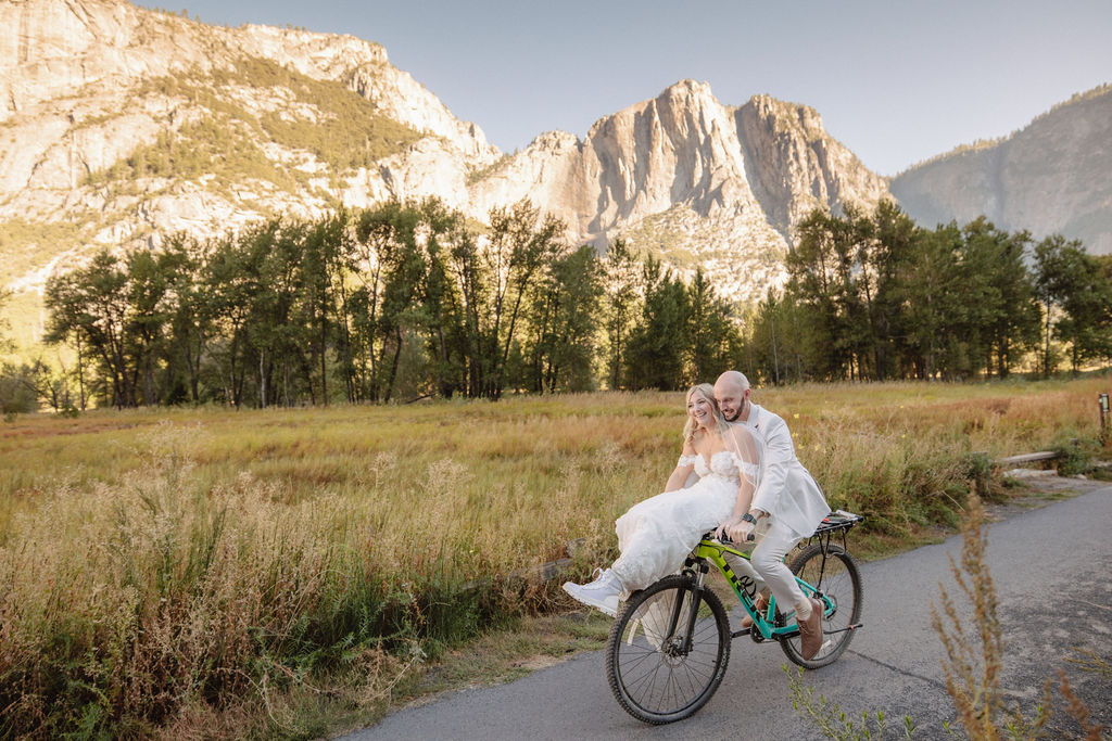 A couple rides a bicycle on a path through a grassy field with tall mountains in the background for their yosemite wedding